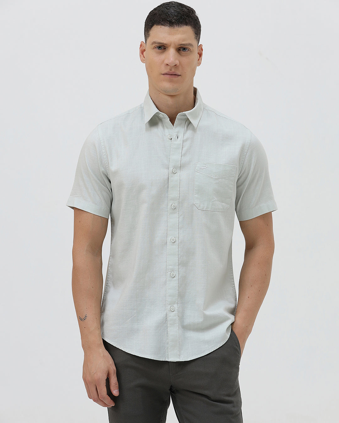 A man wearing a light sage green short-sleeve button-down shirt and dark gray trousers, standing against a plain light gray studio background.