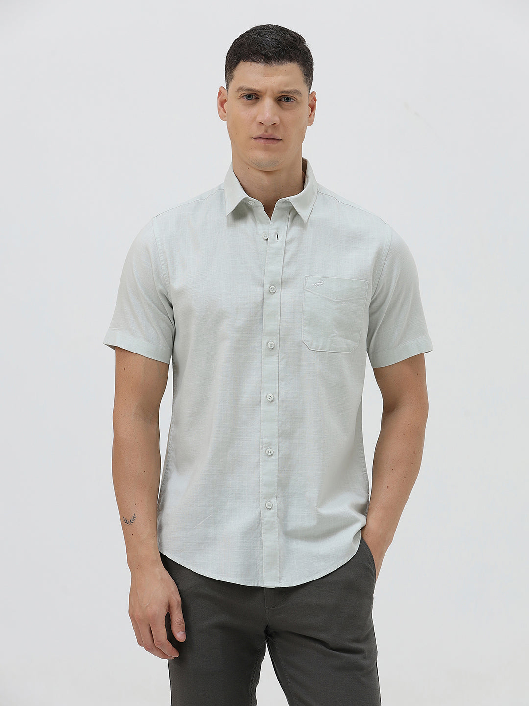 A man wearing a light sage green short-sleeve button-down shirt and dark gray trousers, standing against a plain light gray studio background.