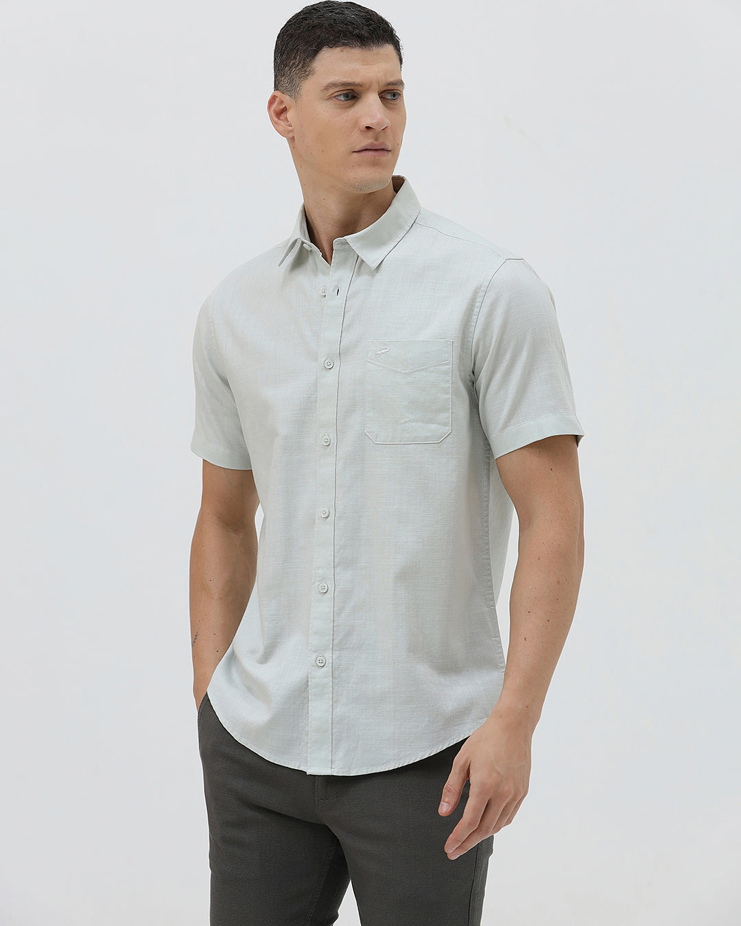 A man wearing a light sage green short-sleeve button-down shirt and dark gray trousers, standing against a plain light gray studio background.