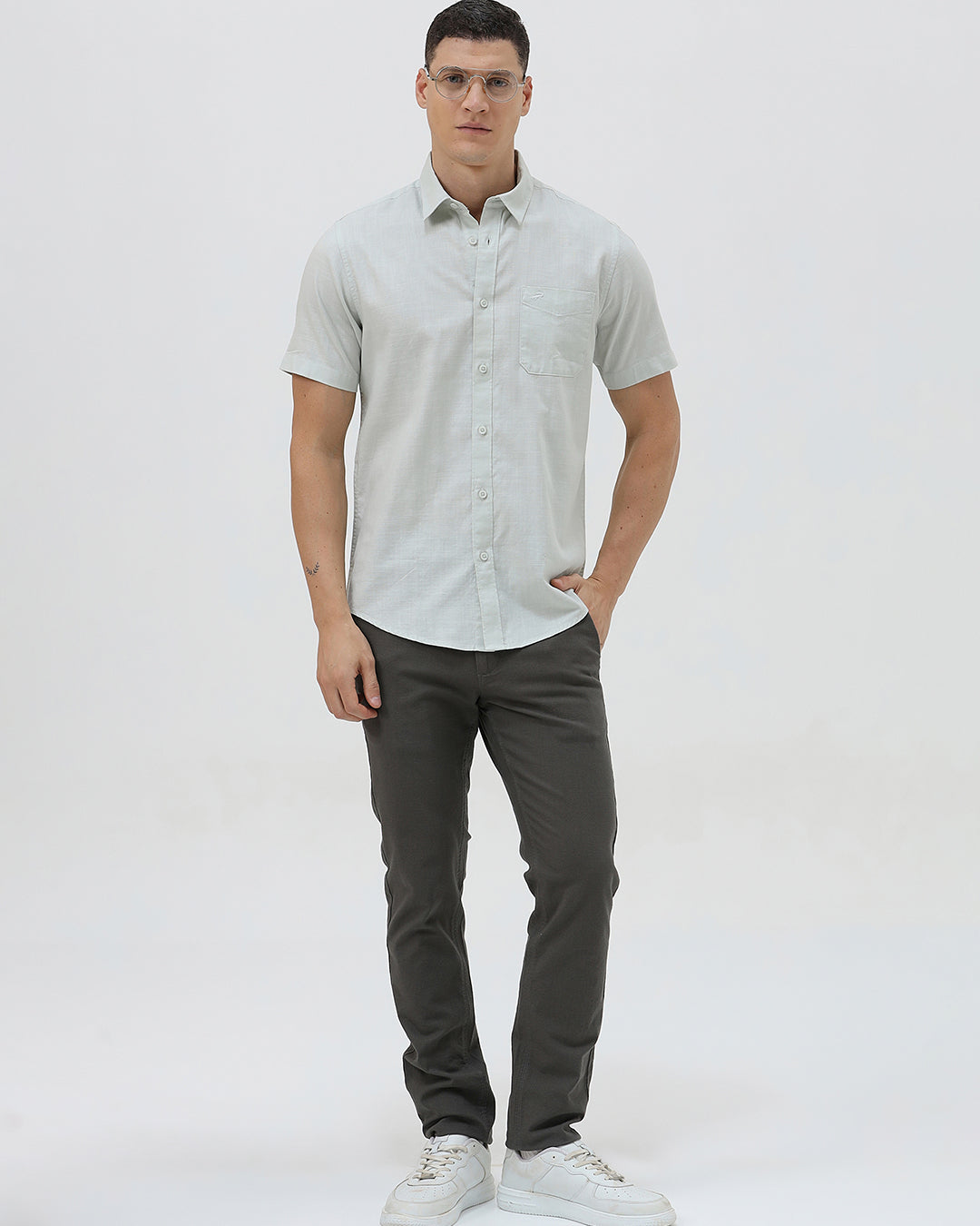 A man wearing a light sage green short-sleeve button-down shirt and dark gray trousers, standing against a plain light gray studio background.