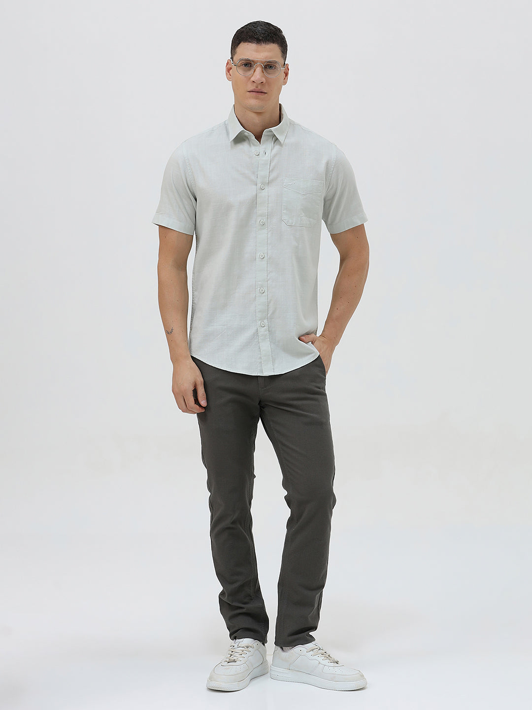 A man wearing a light sage green short-sleeve button-down shirt and dark gray trousers, standing against a plain light gray studio background.
