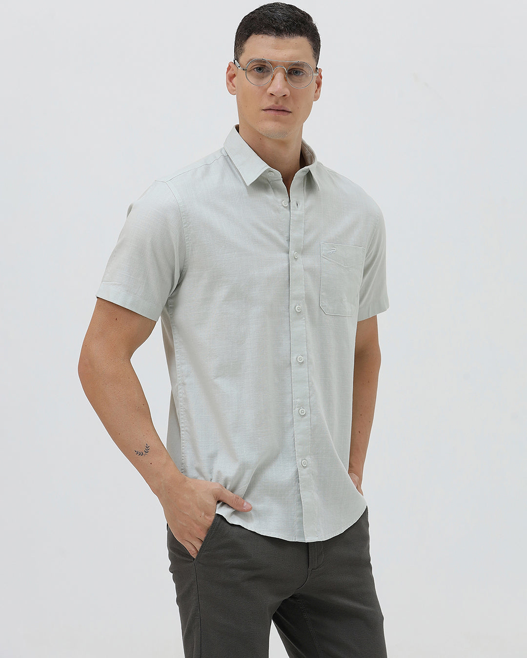 A man wearing a light sage green short-sleeve button-down shirt and dark gray trousers, standing against a plain light gray studio background.
