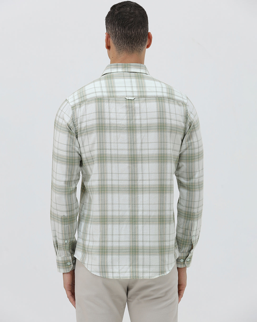 A man wearing a light green and white plaid long-sleeve button-up shirt with beige trousers, standing against a plain light gray background.