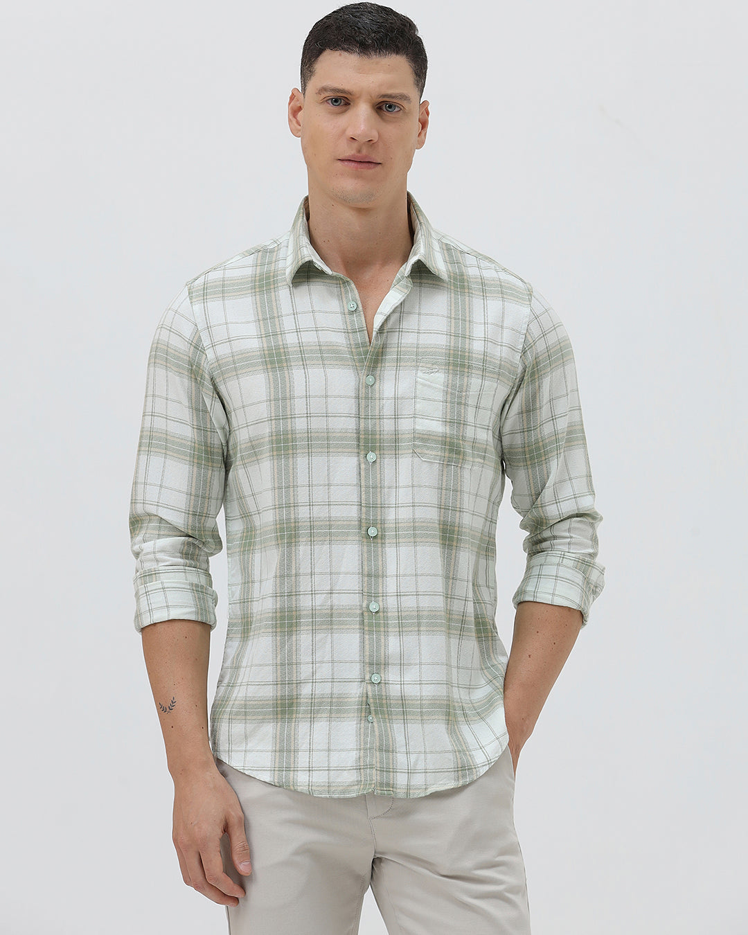 A man wearing a light green and white plaid long-sleeve button-up shirt with beige trousers, standing against a plain light gray background.