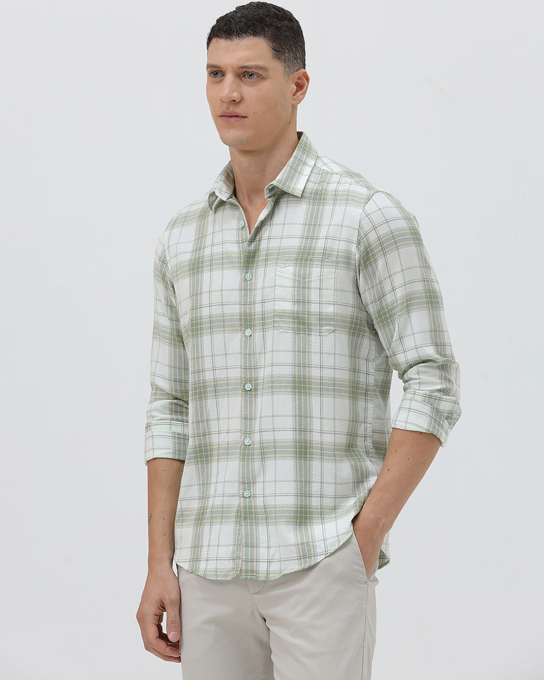 A man wearing a light green and white plaid long-sleeve button-up shirt with beige trousers, standing against a plain light gray background.