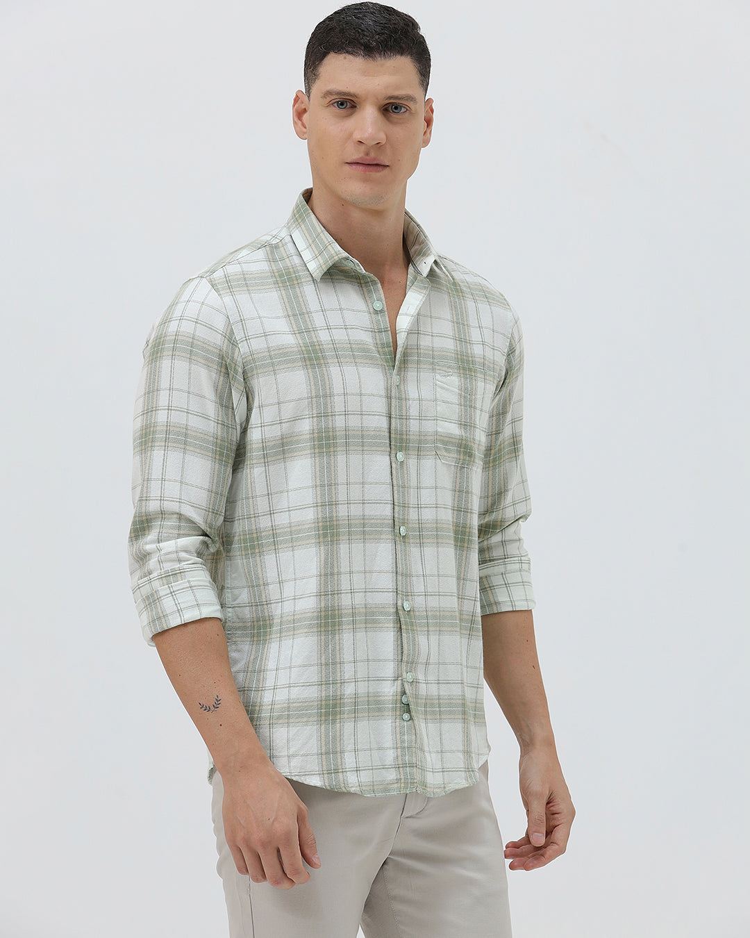 A man wearing a light green and white plaid long-sleeve button-up shirt with beige trousers, standing against a plain light gray background.