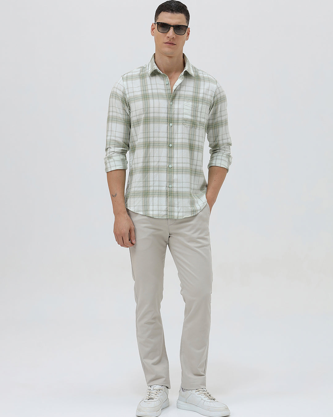 A man wearing a light green and white plaid long-sleeve button-up shirt with beige trousers, standing against a plain light gray background.