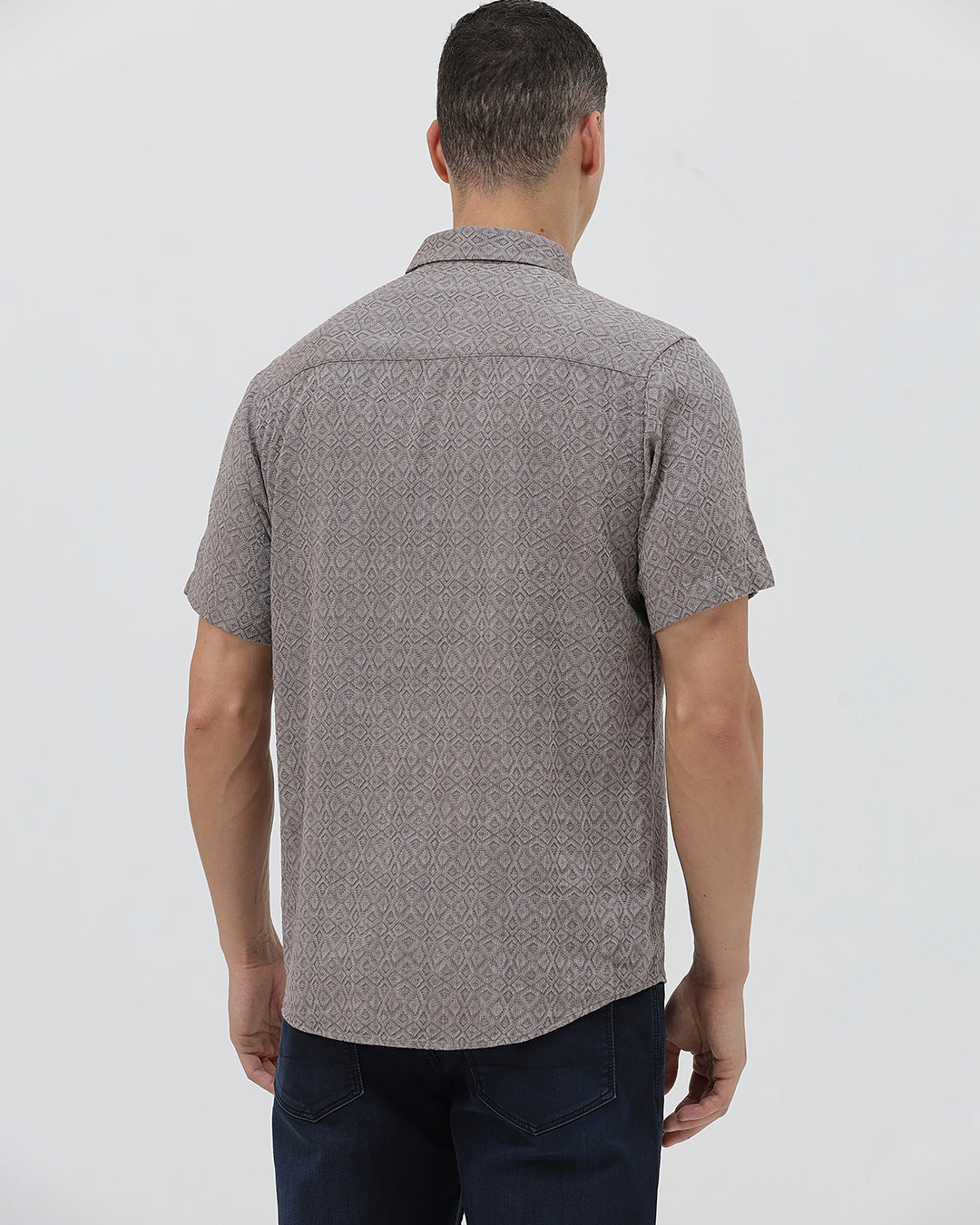 A man wearing a short-sleeve button-up shirt with a fine geometric pattern in gray and brown, paired with dark blue jeans, standing against a light gray studio background.