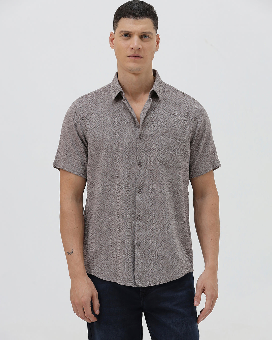 A man wearing a short-sleeve button-up shirt with a fine geometric pattern in gray and brown, paired with dark blue jeans, standing against a light gray studio background.