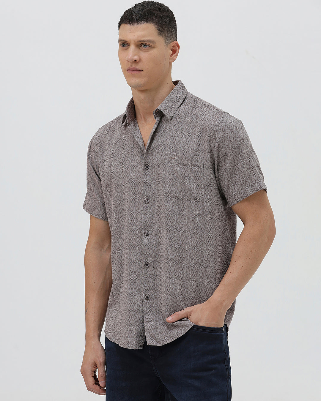 A man wearing a short-sleeve button-up shirt with a fine geometric pattern in gray and brown, paired with dark blue jeans, standing against a light gray studio background.