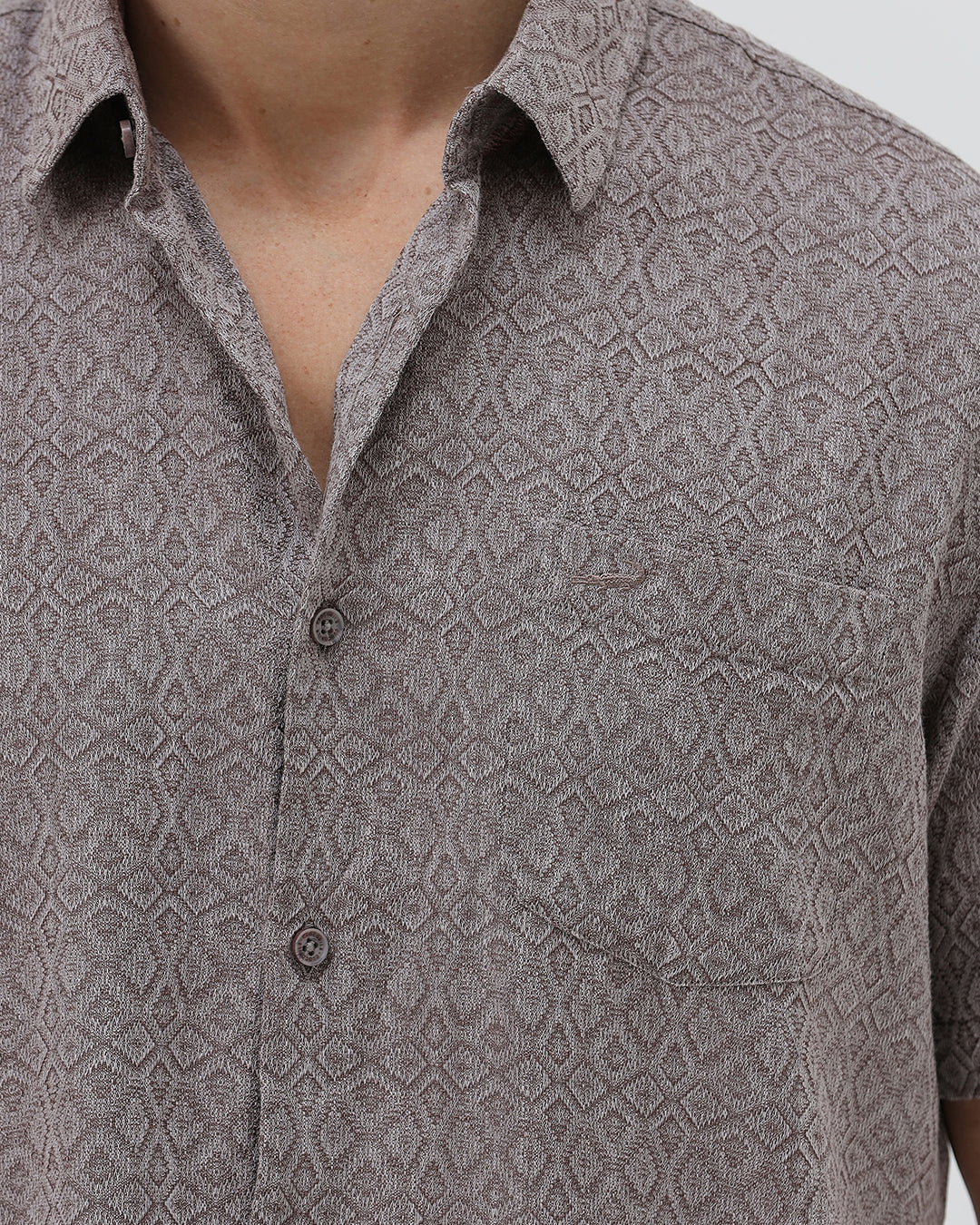 A man wearing a short-sleeve button-up shirt with a fine geometric pattern in gray and brown, paired with dark blue jeans, standing against a light gray studio background.