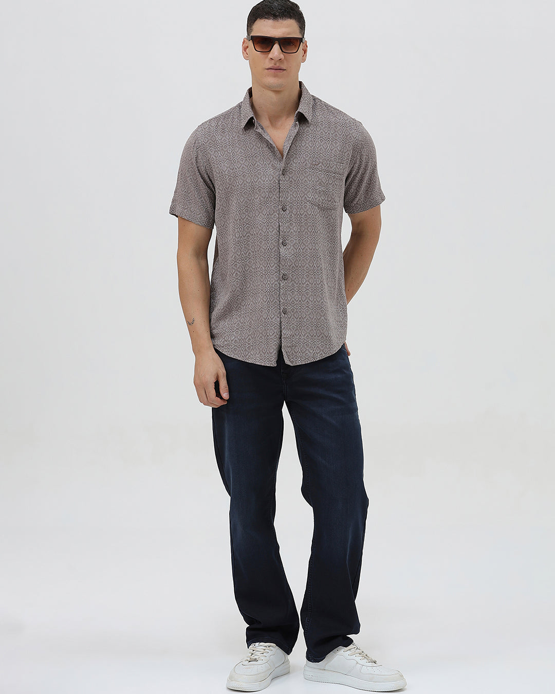 A man wearing a short-sleeve button-up shirt with a fine geometric pattern in gray and brown, paired with dark blue jeans, standing against a light gray studio background.