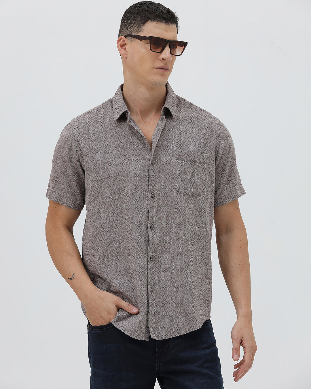 A man wearing a short-sleeve button-up shirt with a fine geometric pattern in gray and brown, paired with dark blue jeans, standing against a light gray studio background.
