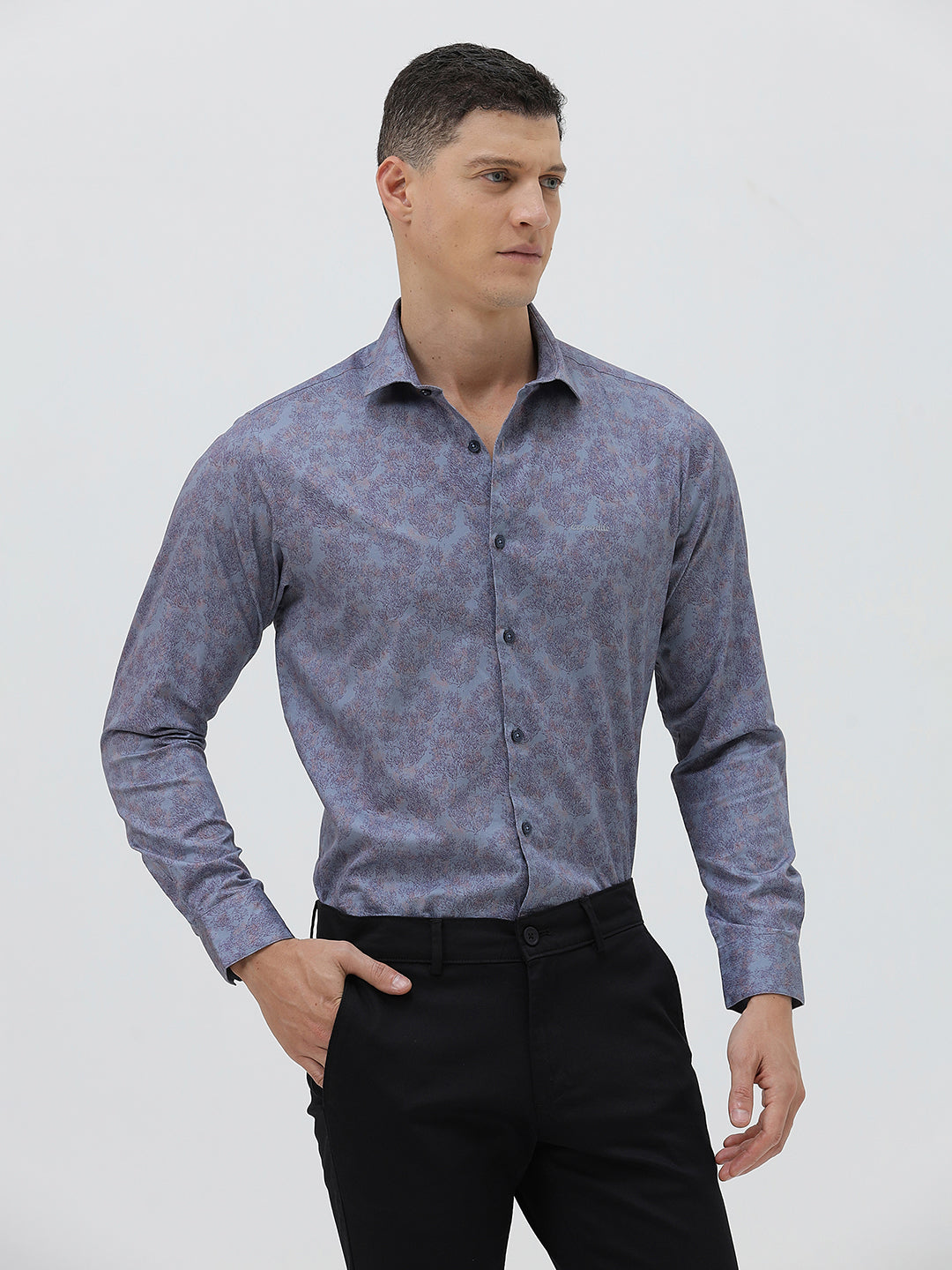 A man wearing a long-sleeve button-up shirt with a subtle blue and purple abstract pattern, paired with black trousers, standing against a light gray studio background.