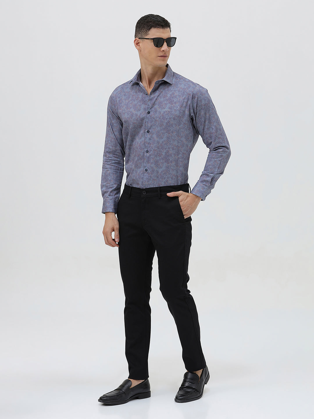 A man wearing a long-sleeve button-up shirt with a subtle blue and purple abstract pattern, paired with black trousers, standing against a light gray studio background.