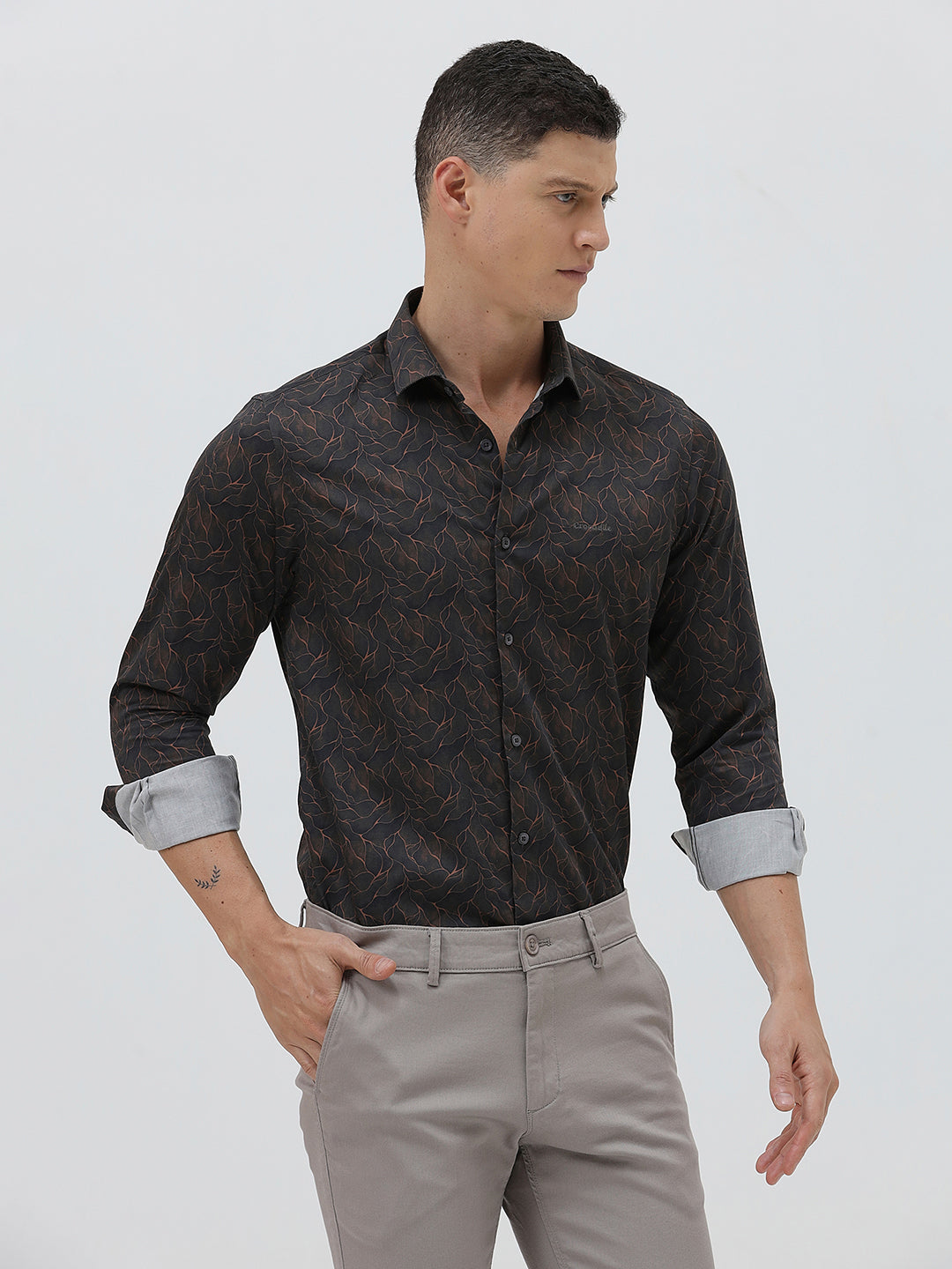 A man wearing a dark brown patterned long-sleeve button-down shirt with rolled-up sleeves and light gray trousers, standing against a light gray studio background.