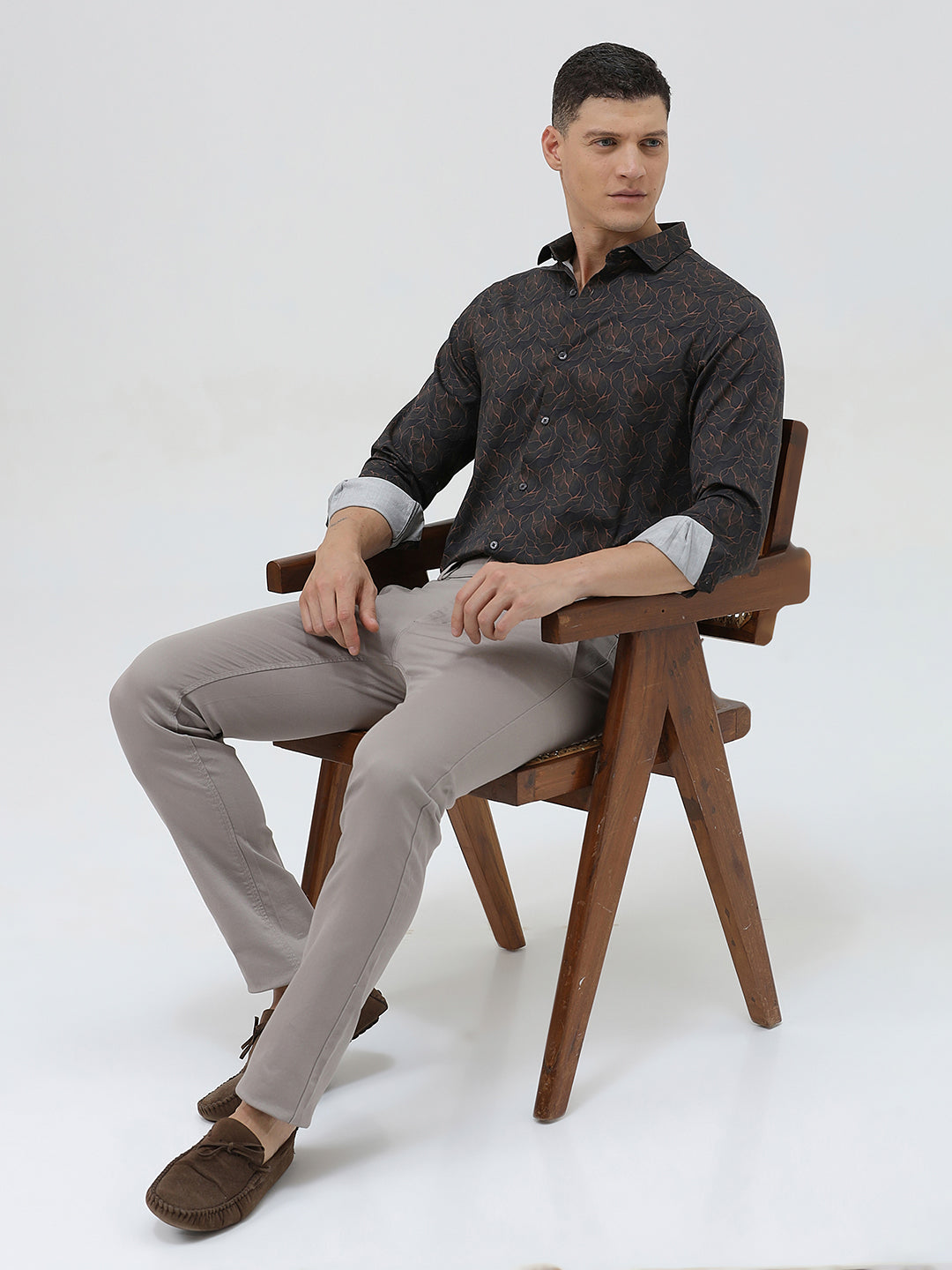 A man wearing a dark brown patterned long-sleeve button-down shirt with rolled-up sleeves and light gray trousers, standing against a light gray studio background.
