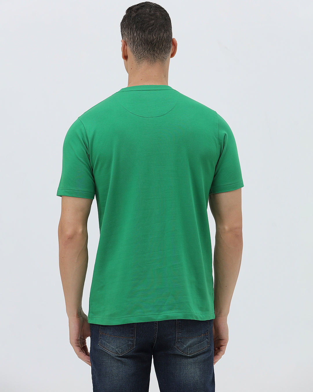 A man wearing a bright green crew-neck t-shirt with a small gold-toned graphic on the chest, paired with dark blue jeans, standing against a light gray studio background.