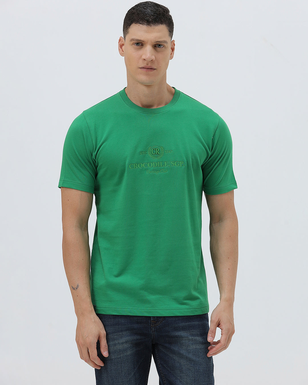 A man wearing a bright green crew-neck t-shirt with a small gold-toned graphic on the chest, paired with dark blue jeans, standing against a light gray studio background.