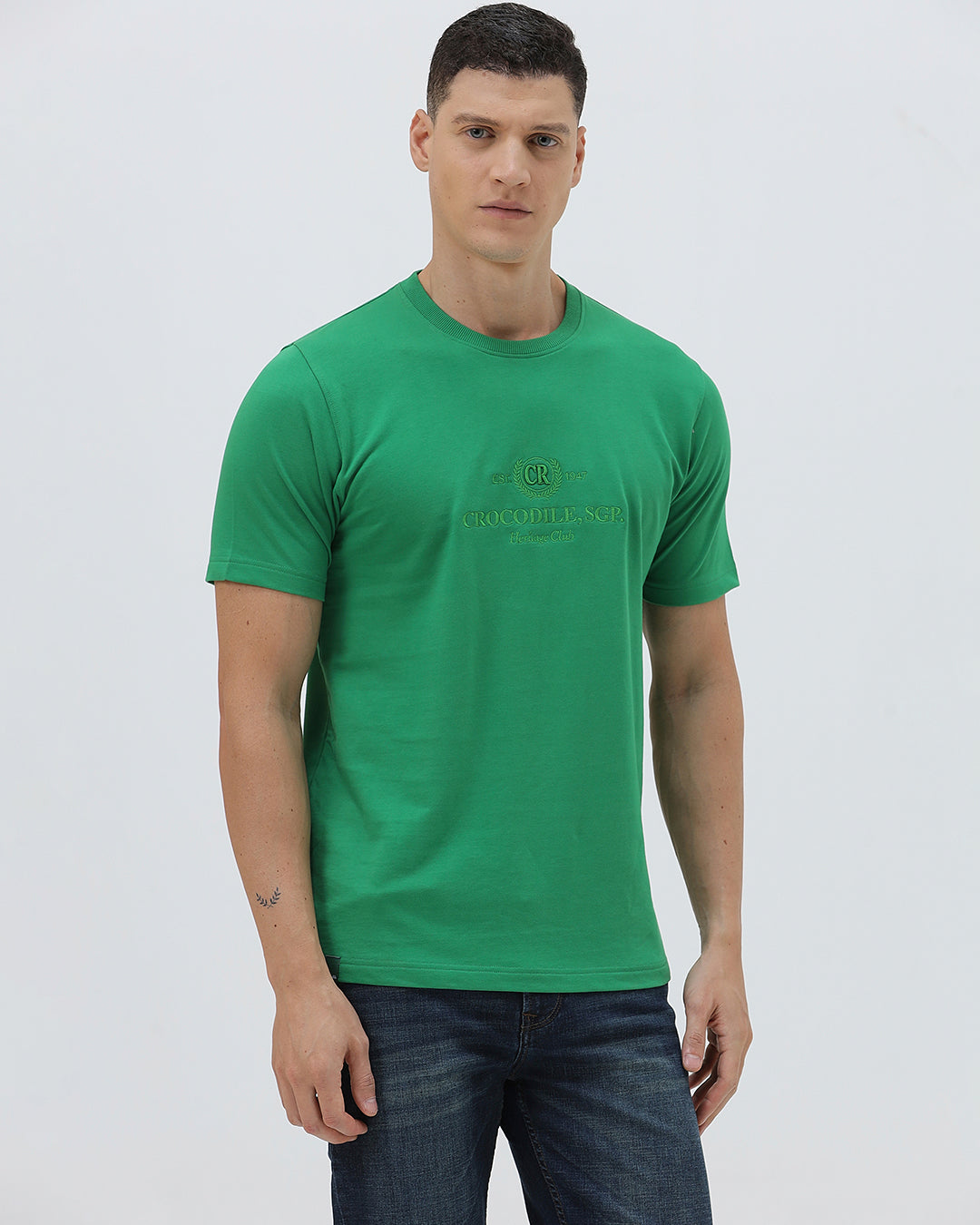 A man wearing a bright green crew-neck t-shirt with a small gold-toned graphic on the chest, paired with dark blue jeans, standing against a light gray studio background.
