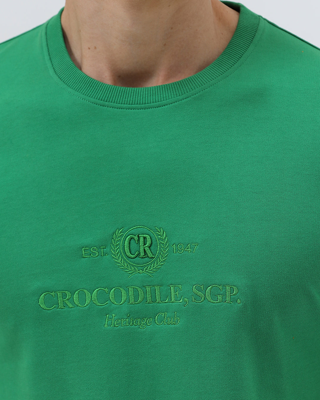 A man wearing a bright green crew-neck t-shirt with a small gold-toned graphic on the chest, paired with dark blue jeans, standing against a light gray studio background.