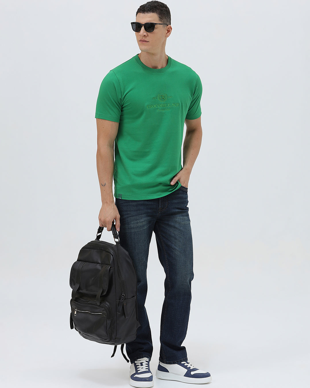 A man wearing a bright green crew-neck t-shirt with a small gold-toned graphic on the chest, paired with dark blue jeans, standing against a light gray studio background.