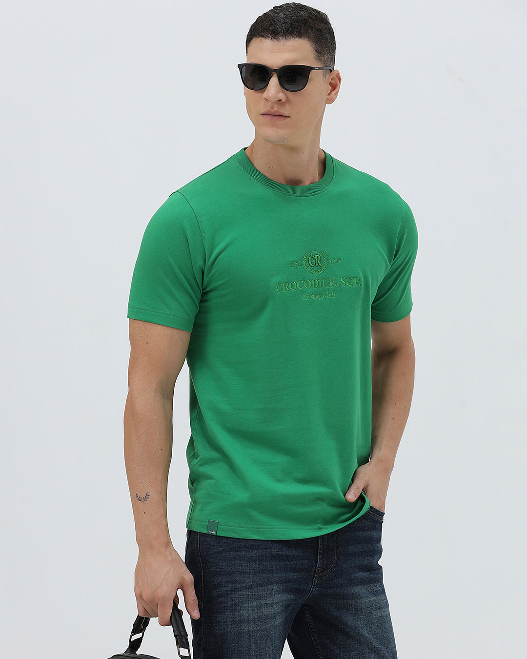A man wearing a bright green crew-neck t-shirt with a small gold-toned graphic on the chest, paired with dark blue jeans, standing against a light gray studio background.