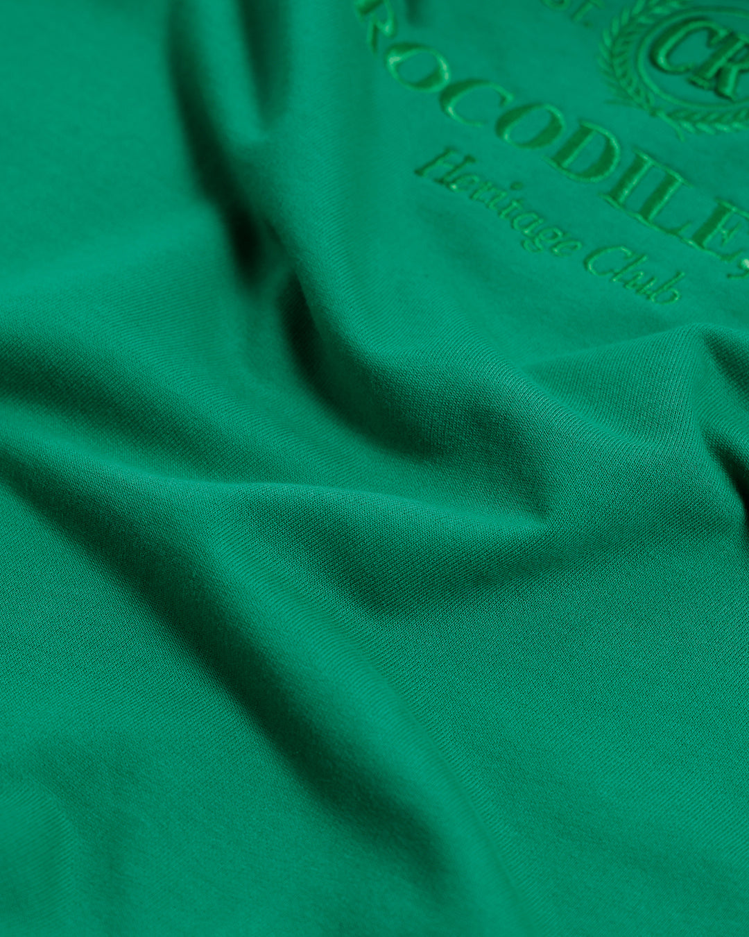 A man wearing a bright green crew-neck t-shirt with a small gold-toned graphic on the chest, paired with dark blue jeans, standing against a light gray studio background.
