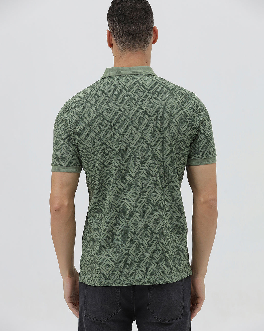 A man wearing a short-sleeve polo shirt with a green and dark gray geometric diamond pattern, paired with dark pants, standing against a light gray studio background.