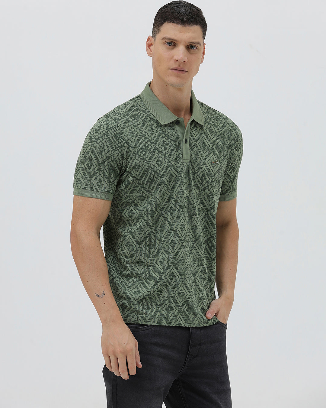 A man wearing a short-sleeve polo shirt with a green and dark gray geometric diamond pattern, paired with dark pants, standing against a light gray studio background.