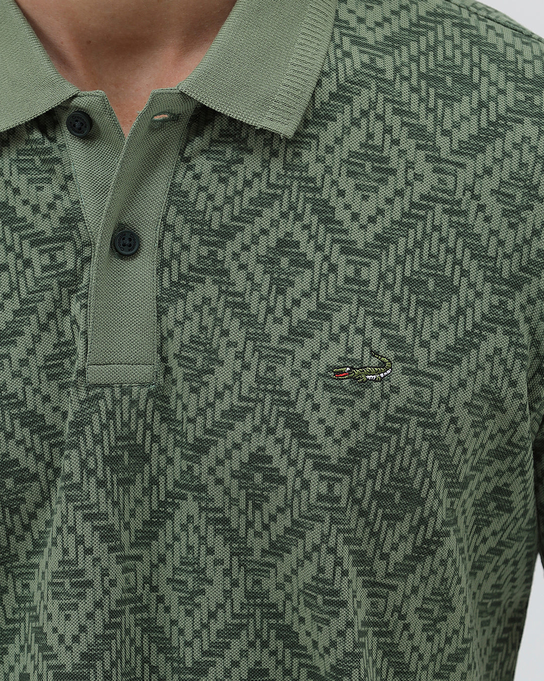 A man wearing a short-sleeve polo shirt with a green and dark gray geometric diamond pattern, paired with dark pants, standing against a light gray studio background.