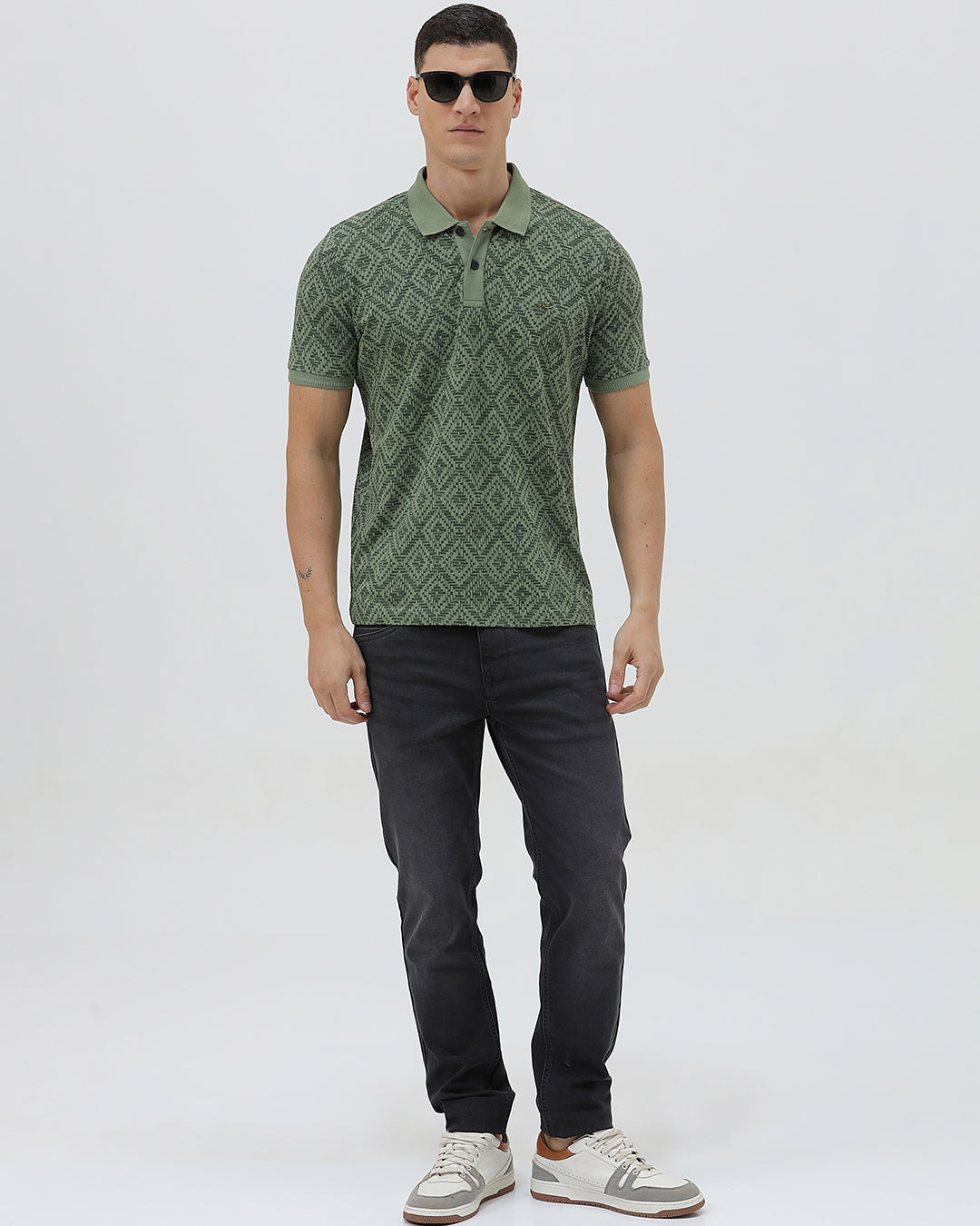 A man wearing a short-sleeve polo shirt with a green and dark gray geometric diamond pattern, paired with dark pants, standing against a light gray studio background.