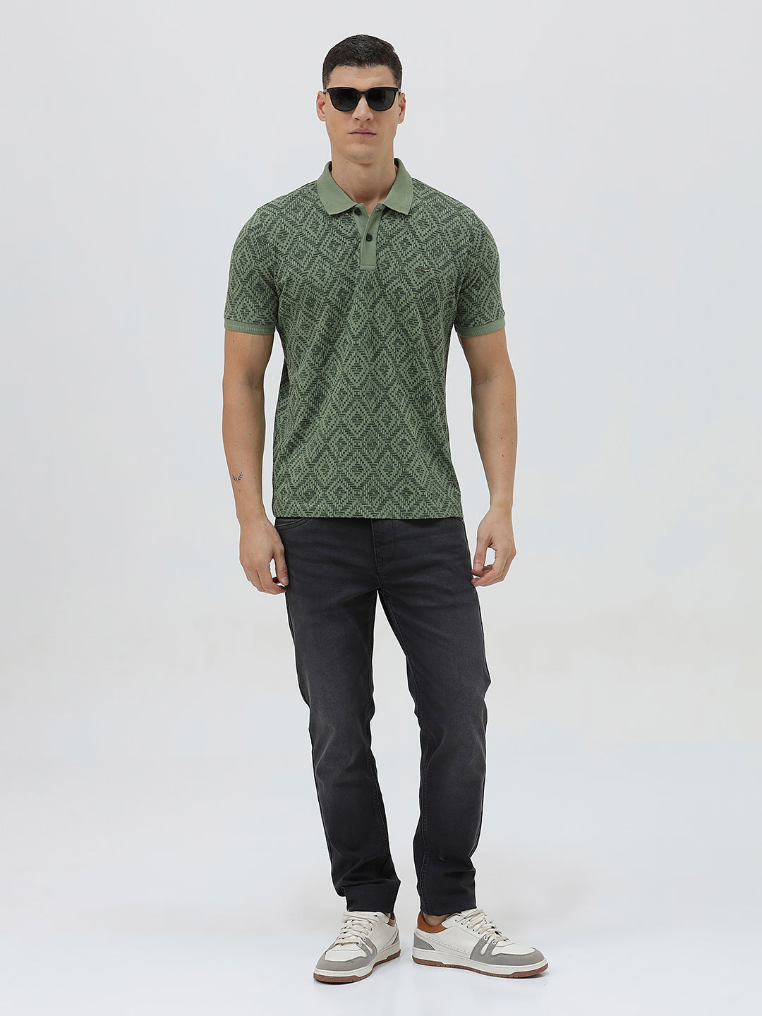 A man wearing a short-sleeve polo shirt with a green and dark gray geometric diamond pattern, paired with dark pants, standing against a light gray studio background.