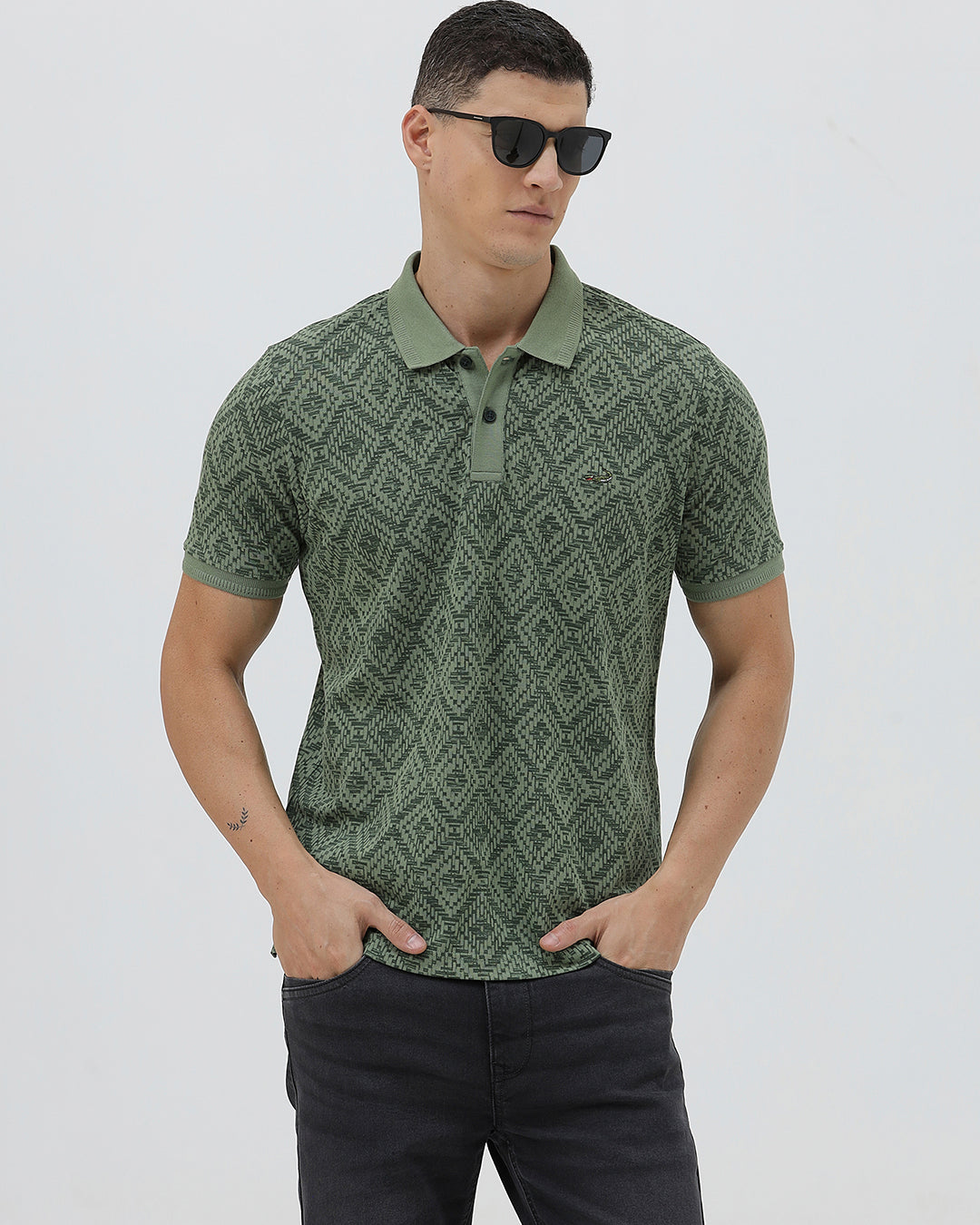 A man wearing a short-sleeve polo shirt with a green and dark gray geometric diamond pattern, paired with dark pants, standing against a light gray studio background.