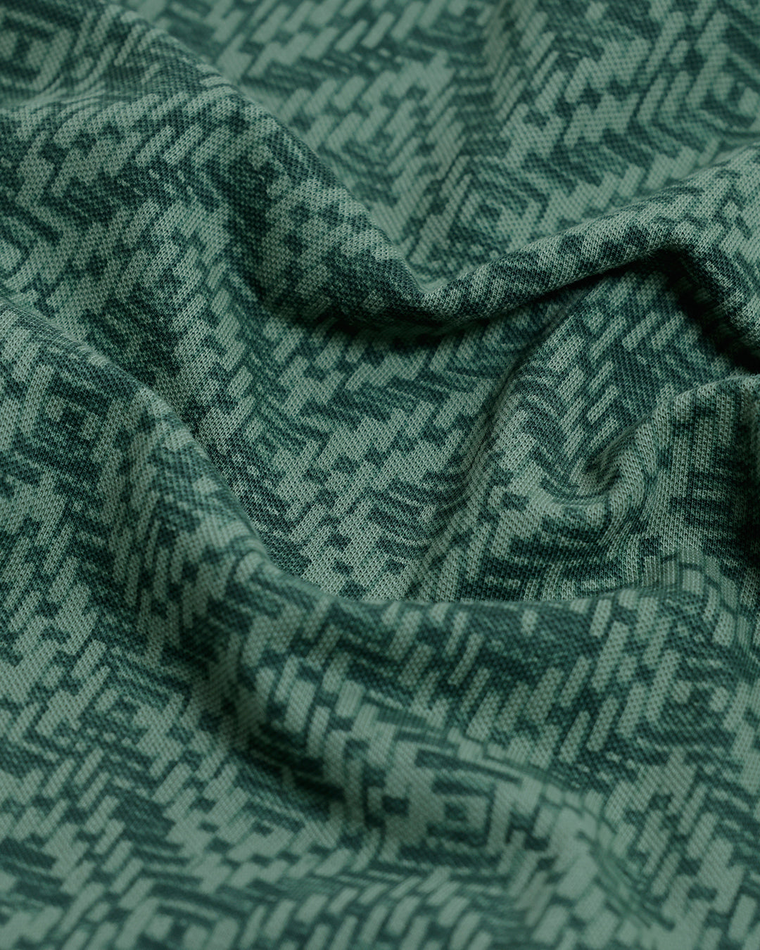 A man wearing a short-sleeve polo shirt with a green and dark gray geometric diamond pattern, paired with dark pants, standing against a light gray studio background.