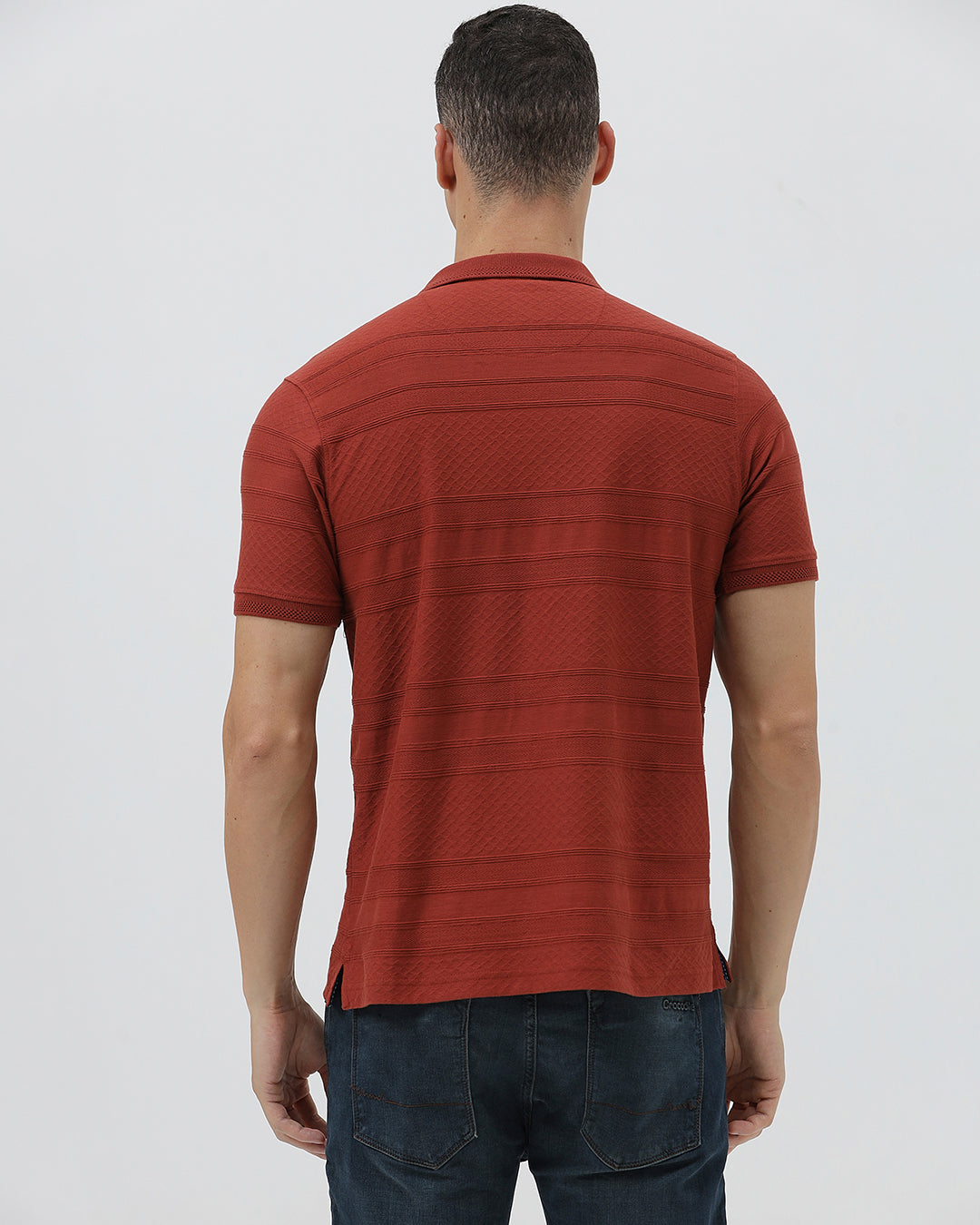 A man wearing a rust-red short-sleeve polo shirt with a subtle textured pattern and dark blue jeans, standing against a light gray studio background.
