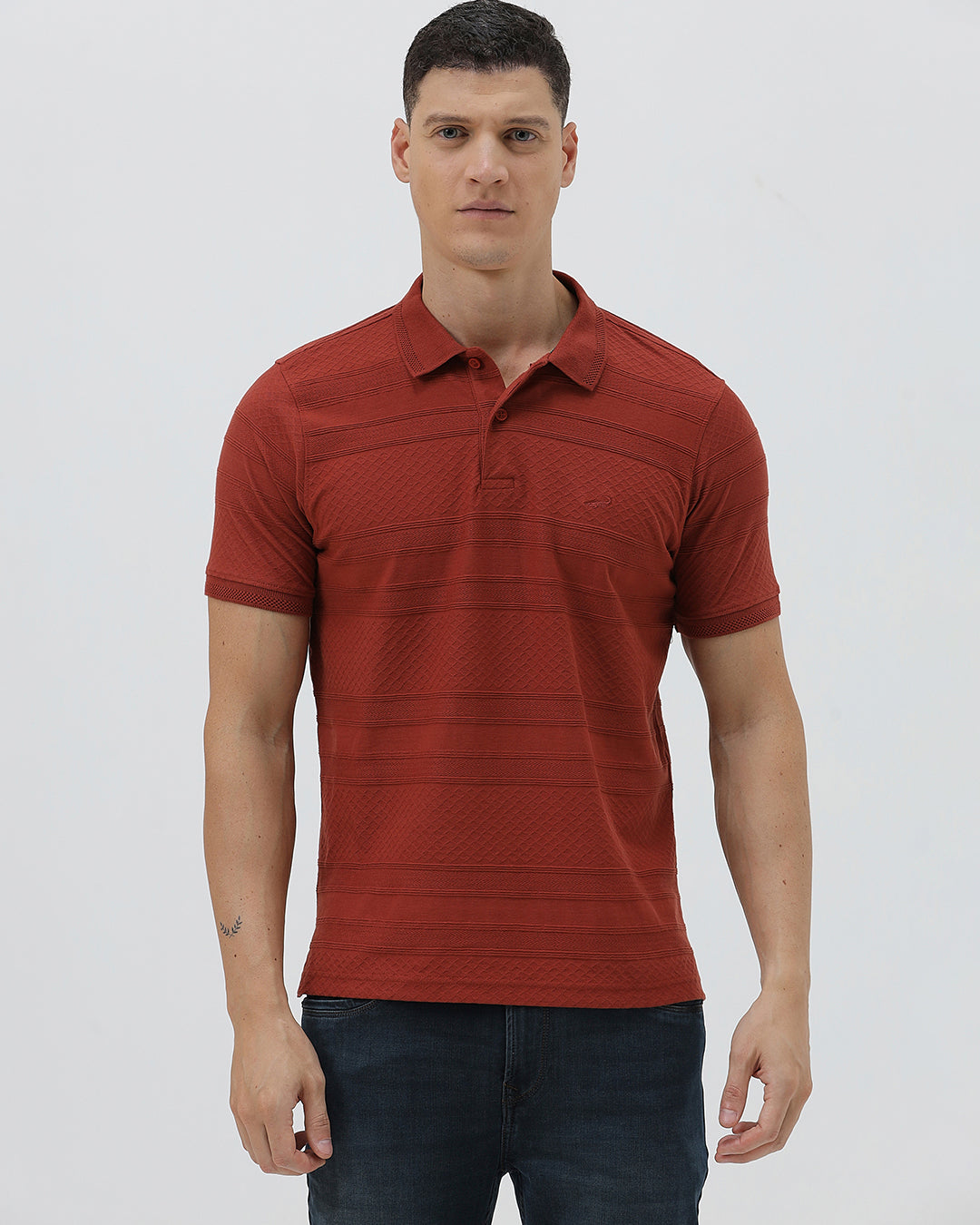 A man wearing a rust-red short-sleeve polo shirt with a subtle textured pattern and dark blue jeans, standing against a light gray studio background.