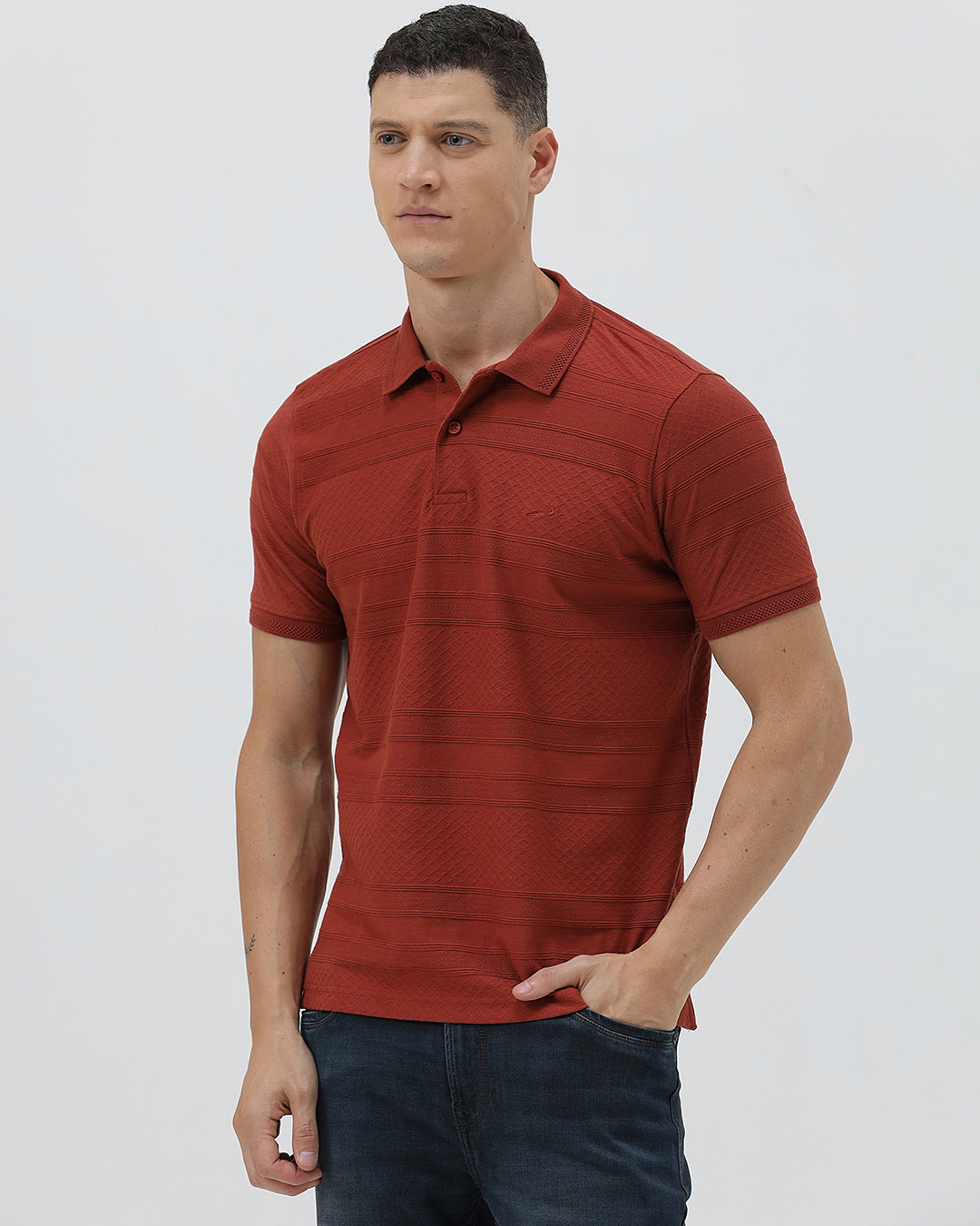 A man wearing a rust-red short-sleeve polo shirt with a subtle textured pattern and dark blue jeans, standing against a light gray studio background.