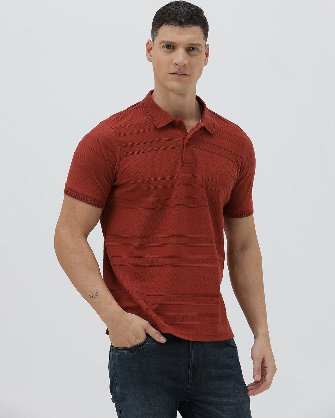 A man wearing a rust-red short-sleeve polo shirt with a subtle textured pattern and dark blue jeans, standing against a light gray studio background.