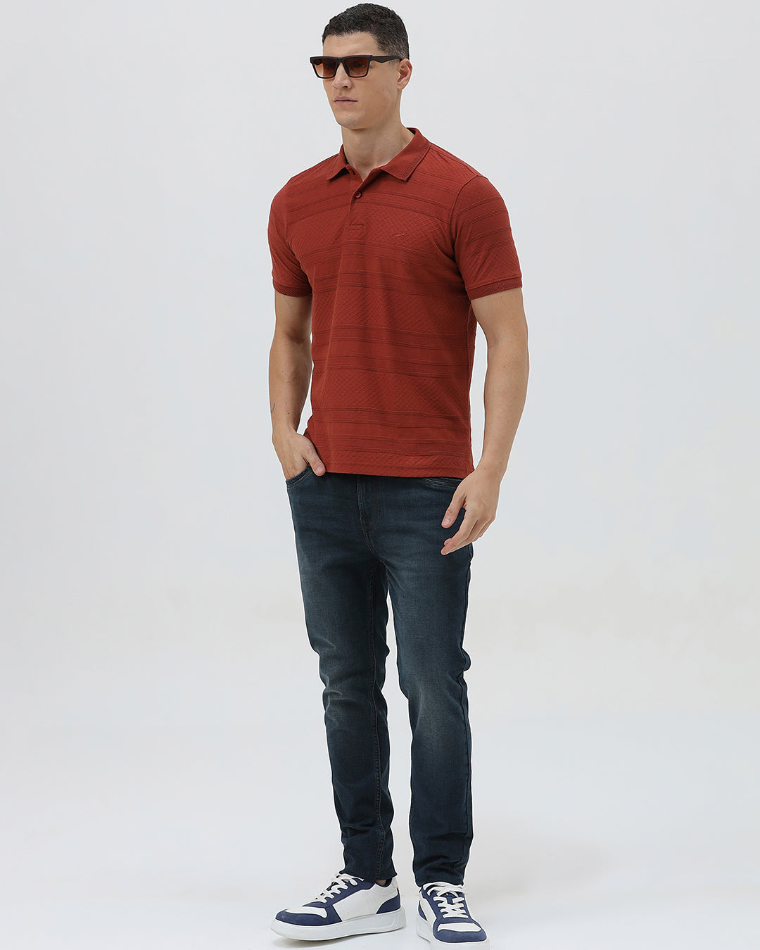 A man wearing a rust-red short-sleeve polo shirt with a subtle textured pattern and dark blue jeans, standing against a light gray studio background.