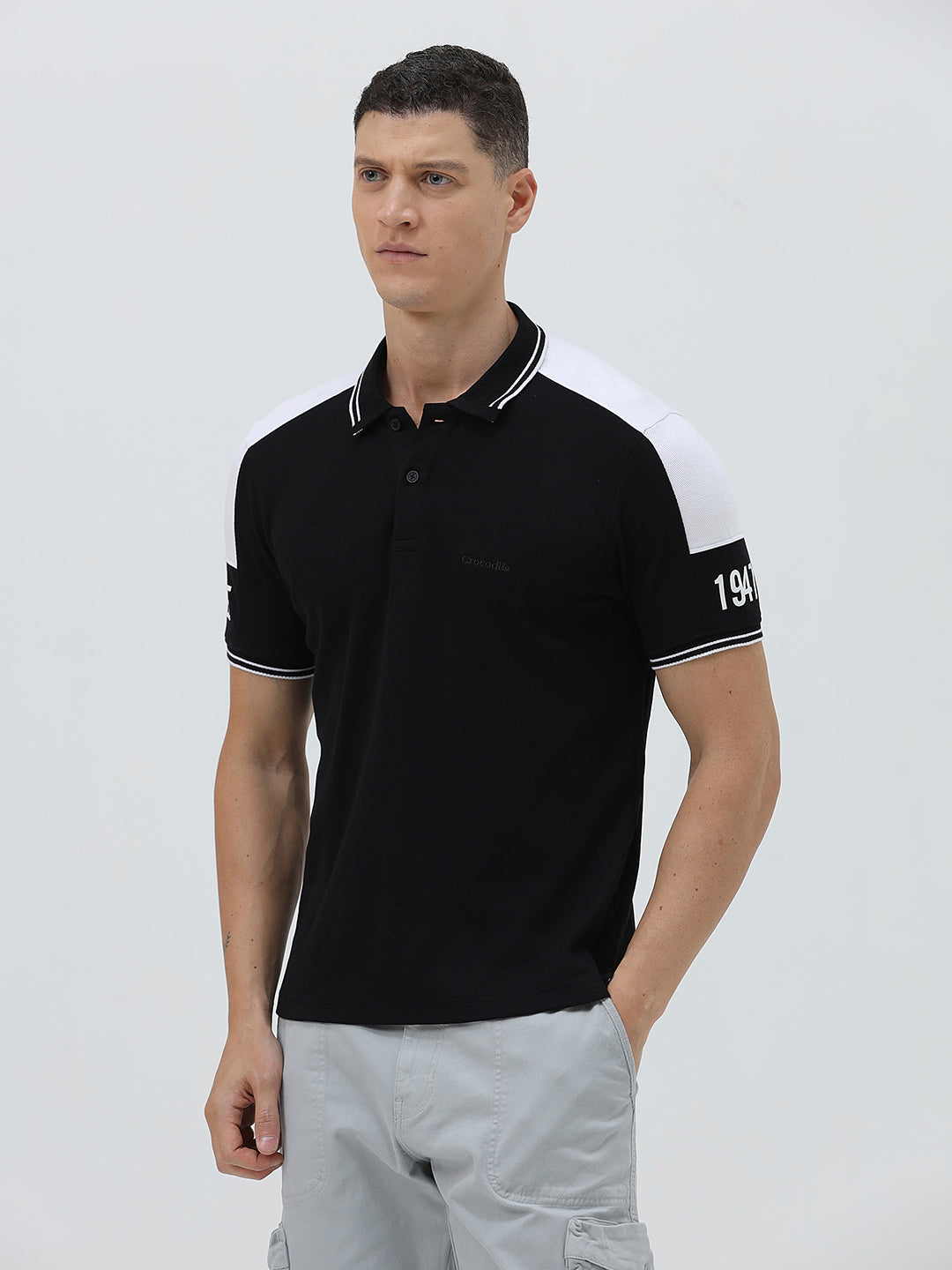 A man modeling a black short-sleeve polo shirt with white shoulder panels and white striped trim on the collar and sleeve cuffs, paired with light gray cargo pants against a plain light gray background.