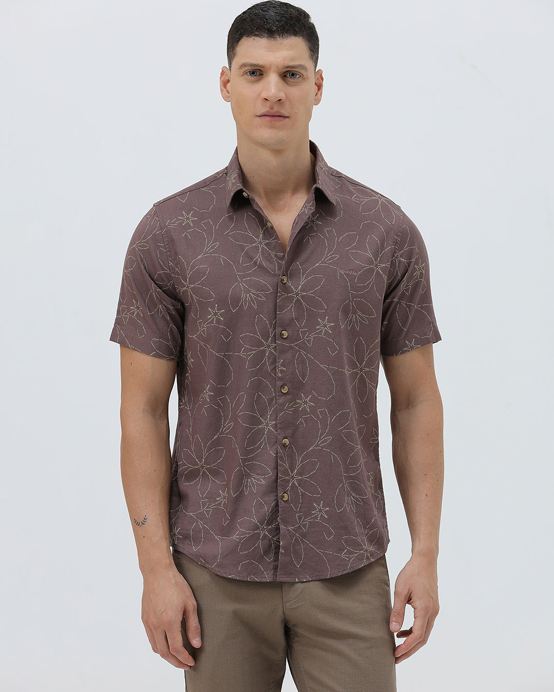 A man wearing a short-sleeve button-up shirt in a muted brownish-purple color with a light beige floral pattern, paired with khaki pants, against a plain light gray background.
