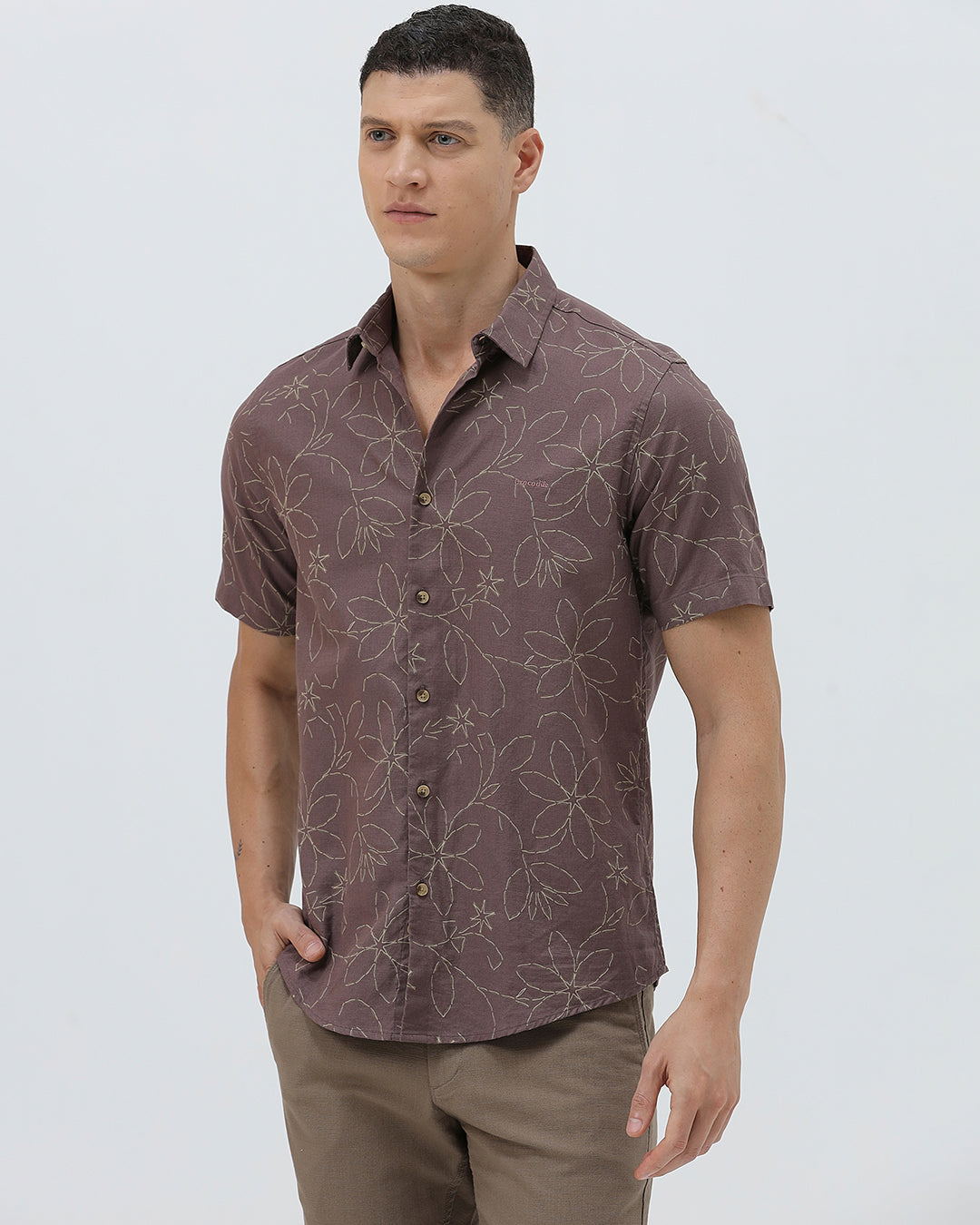 A man wearing a short-sleeve button-up shirt in a muted brownish-purple color with a light beige floral pattern, paired with khaki pants, against a plain light gray background.