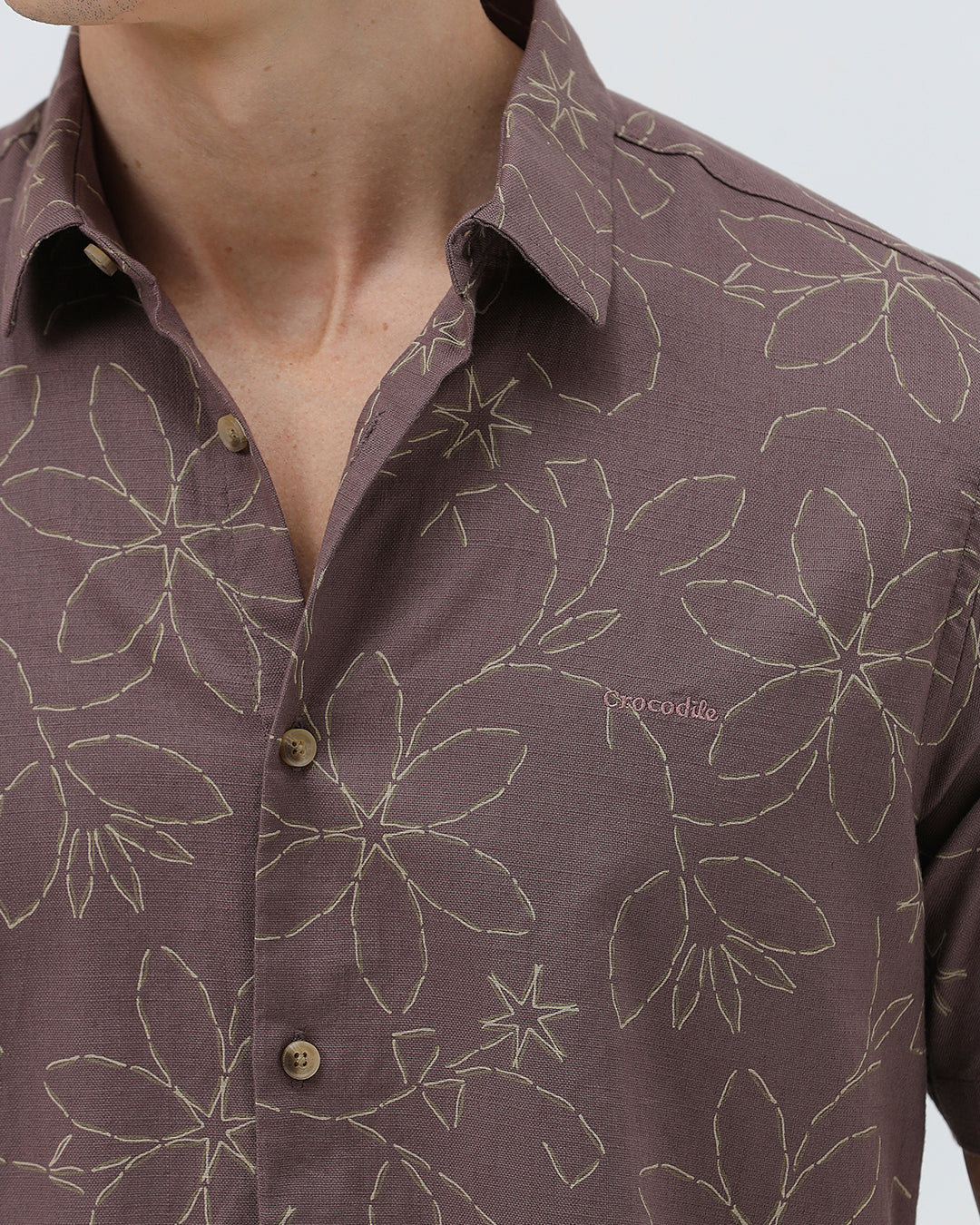 A man wearing a short-sleeve button-up shirt in a muted brownish-purple color with a light beige floral pattern, paired with khaki pants, against a plain light gray background.