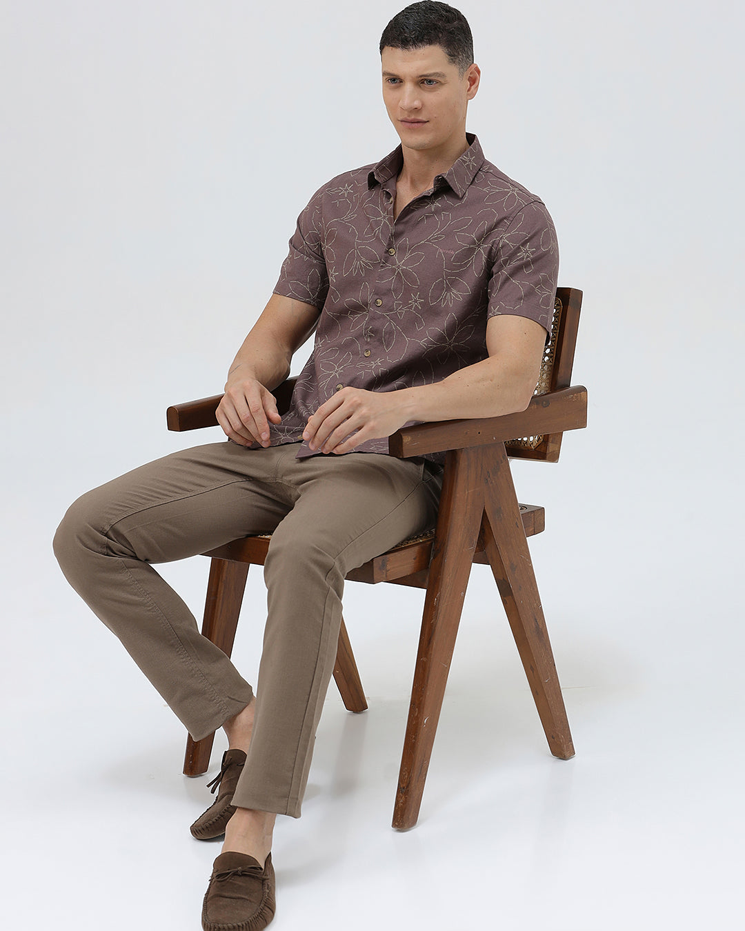 A man wearing a short-sleeve button-up shirt in a muted brownish-purple color with a light beige floral pattern, paired with khaki pants, against a plain light gray background.