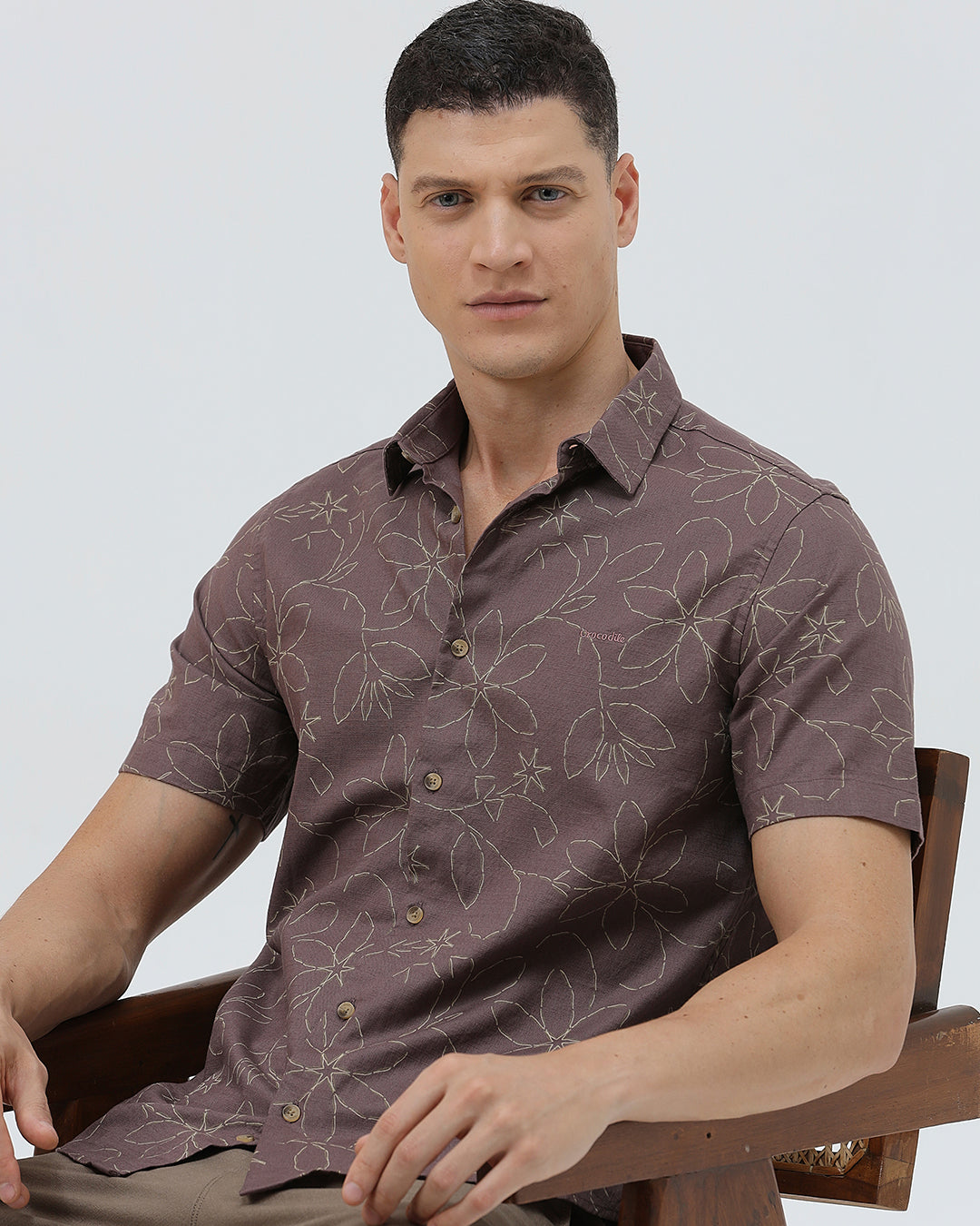 A man wearing a short-sleeve button-up shirt in a muted brownish-purple color with a light beige floral pattern, paired with khaki pants, against a plain light gray background.