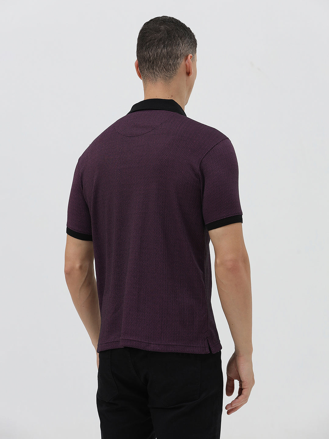 A man wearing a short-sleeve polo shirt in dark plum purple with a black collar and black sleeve trim, paired with black pants, against a plain light gray background.