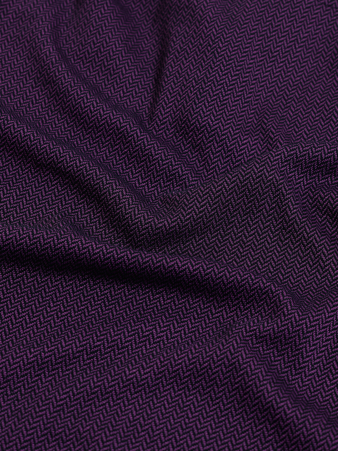 A man wearing a short-sleeve polo shirt in dark plum purple with a black collar and black sleeve trim, paired with black pants, against a plain light gray background.