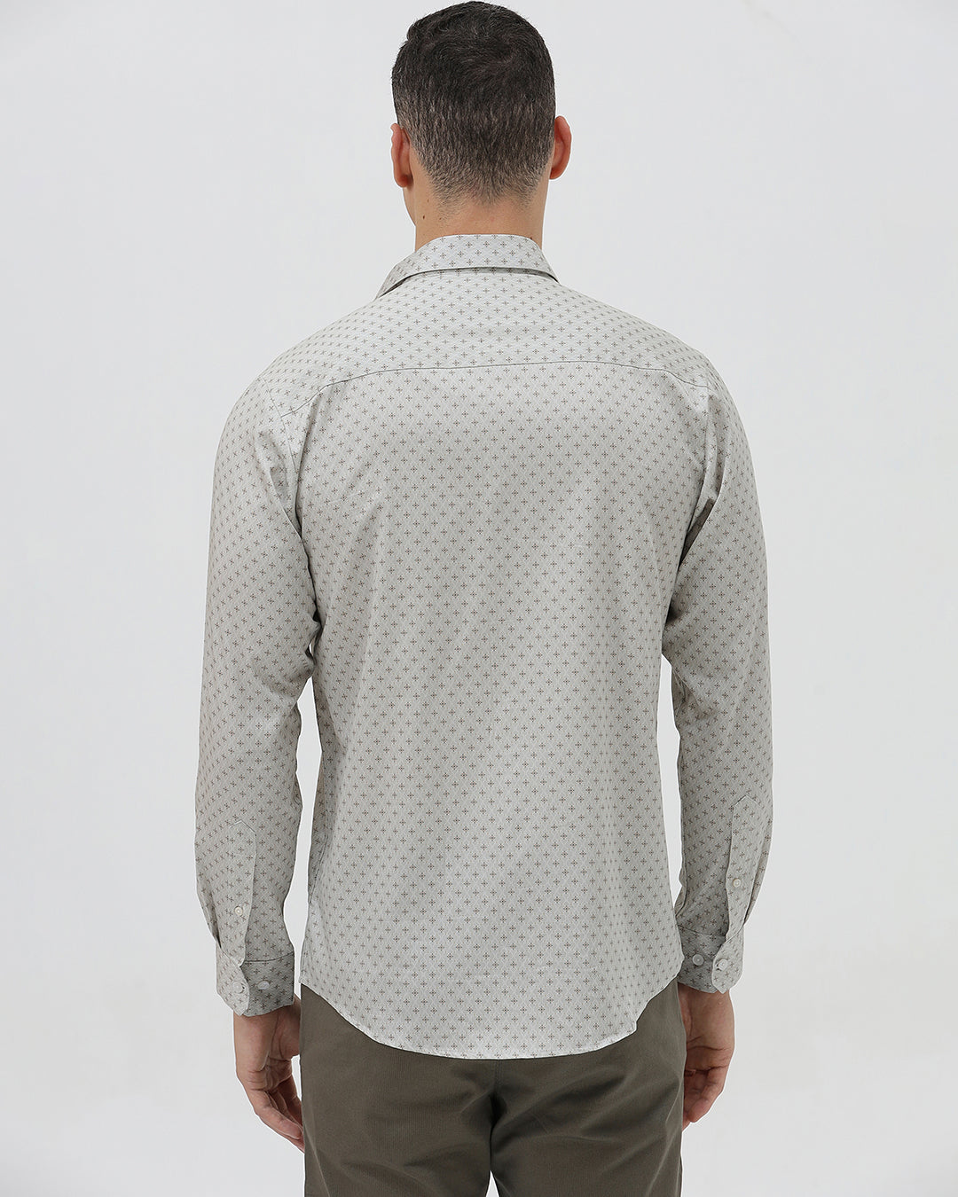 A man wearing a light gray long-sleeve button-down shirt with a small repeating geometric pattern, paired with olive green trousers, against a plain light gray background.