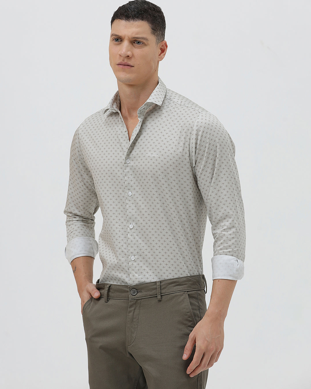 A man wearing a light gray long-sleeve button-down shirt with a small repeating geometric pattern, paired with olive green trousers, against a plain light gray background.